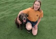 A woman kneeling on the grass with a brown dog.