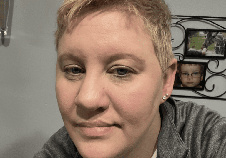 A woman in a gray jacket with blond hair in front of a clock.