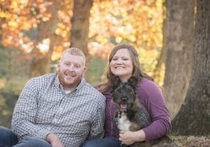 A couple sits on the ground with their dog in the fall.