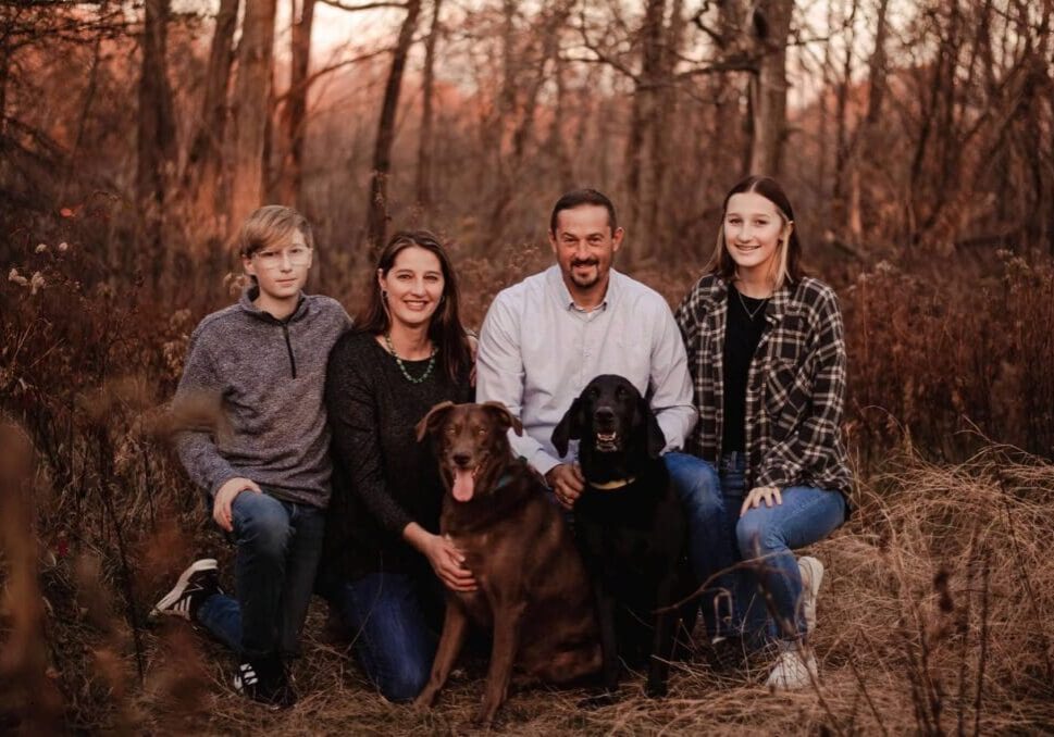 A family poses with their dog in the woods at sunset.